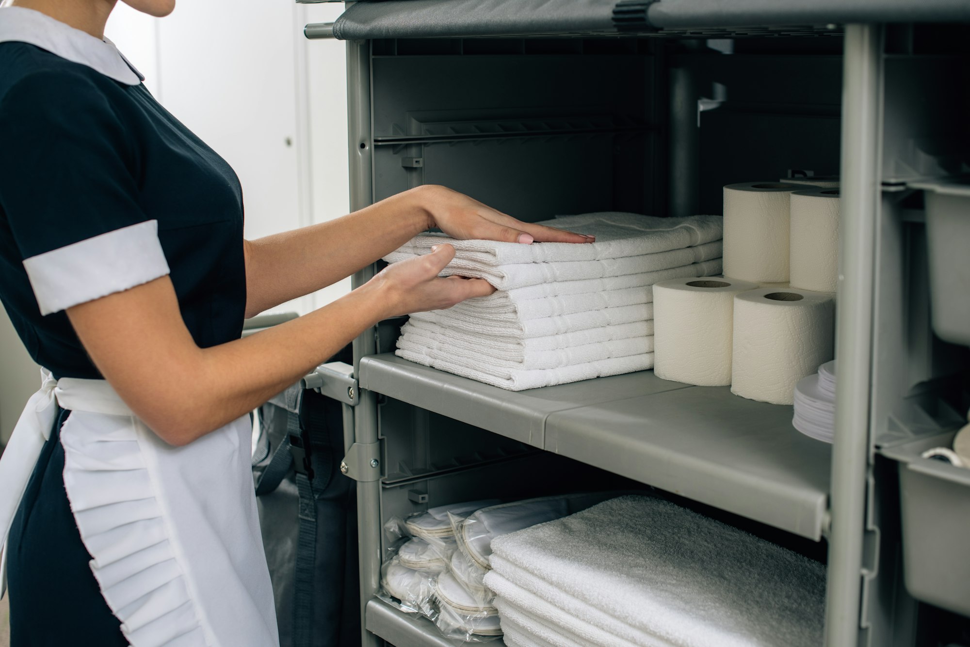 cropped shot of maid in uniform taking towels from shelf of housekeeping cart