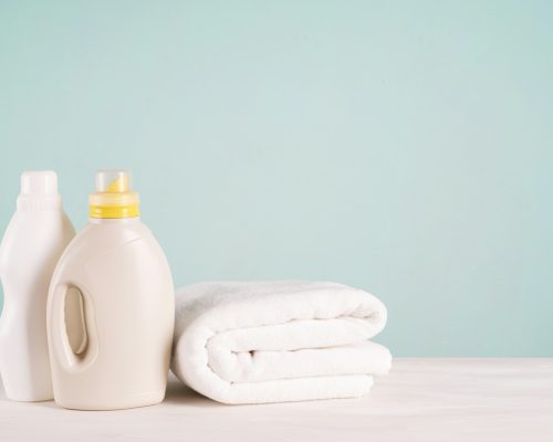 A bottle of laundry detergent, fabric softener and a stack of clean laundry on a blue background.